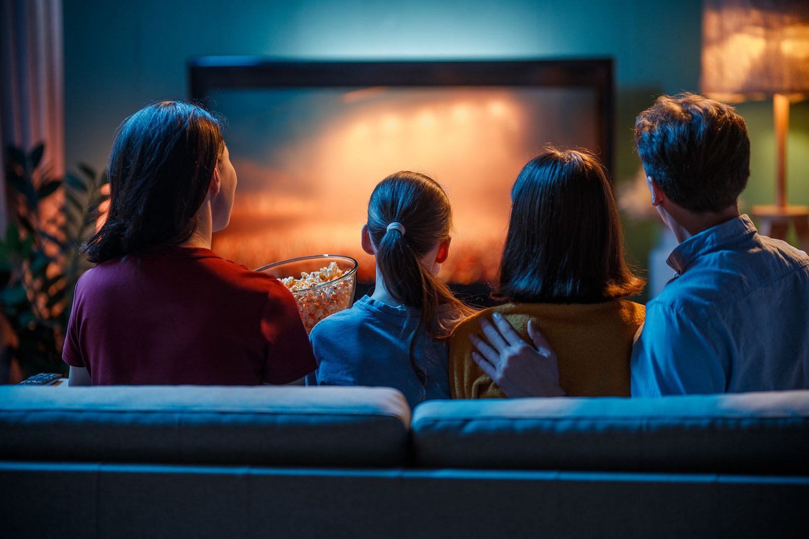 Une famille réunie sur un canapé regarde un programme sur un grand téléviseur, le soir dans un salon